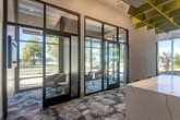 Located at 1240 La Avenida St in Mountain View, California, this contemporary commercial lobby delivers a polished and welcoming first impression through thoughtful architectural design and high-end finishes. The single-floor space is defined by soaring ceilings that amplify the sense of openness, while expansive floor-to-ceiling glass walls and black-framed curtain wall systems flood the interior with natural light and offer unobstructed views of the surrounding landscaped exterior.

A standout centerpiece is the elongated white marble-veined island, which serves as both a functional counter and a bold design statement. Overhead, a sculptural geometric ceiling installation in vibrant chartreuse yellow adds an artistic focal point and contributes to the space's acoustic performance. The patterned carpet flooring in soft blue-grey tones with an organic, fluid design grounds the space with texture and visual interest.

Along the perimeter, built-in cabinetry with a light wood finish provides practical storage, while a cushioned green banquette bench offers a comfortable seating area beside the glazed walls. Glass-enclosed alcoves near the entrance provide semi-private seating nooks with modern lounge chairs. The overall ambiance is bright, contemporary, and professional, making it well-suited for a leasing office, corporate reception, or community hub.