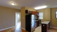 This single-floor residence offers a practical and comfortable layout designed for everyday living. The kitchen serves as a central feature of the home, equipped with stainless steel appliances including a full-size refrigerator, a gas range with oven, and a dishwasher. Rich, dark wood cabinetry provides ample storage overhead and below, complemented by light stone-patterned countertops that add a clean, neutral contrast. The kitchen floor is finished in light tile, which transitions seamlessly into warm hardwood flooring that extends throughout the adjacent living areas. The open sightlines from the kitchen into the rest of the home create a connected and airy feel, with natural light filtering in through windows fitted with blinds. The warm beige wall tones throughout contribute to a welcoming ambiance. A hallway visible from the kitchen leads to additional rooms, hinting at a well-organized floor plan. The arched window detail at the front entry adds a subtle architectural touch, enhancing the overall character of the space. The home combines functional design with comfortable finishes, making it a practical and inviting living environment.
