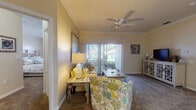 This single-story residence is designed with a cohesive coastal aesthetic carried throughout its rooms. The living room serves as the heart of the home, featuring crown molding, a ceiling fan, and plush carpeting. A sliding glass door floods the space with natural light and provides direct access to an outdoor patio, offering views of surrounding greenery. Coastal-inspired décor, including nautical accents and ocean-toned artwork, reinforces the relaxed atmosphere.

The kitchen is thoughtfully laid out with white cabinetry, a decorative wine rack, and a peninsula with a built-in sink that opens toward the dining area. Black appliances, including a refrigerator, range, and microwave, provide contrast against the light countertops and tile flooring. Adjacent to the kitchen, a dedicated laundry area houses a full-size washer and dryer.

Two bedrooms each feature carpeted floors and large windows with vertical blinds that allow ample natural light while maintaining privacy. A hallway connecting the bedrooms leads to a bathroom appointed with a vanity, tiled flooring, and a full soaking tub with a shower. A walk-in closet is also accessible from the hallway, adding convenient storage throughout the unit.