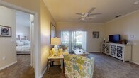 This single-story residence is designed with a cohesive coastal aesthetic carried throughout its rooms. The living room serves as the heart of the home, featuring crown molding, a ceiling fan, and plush carpeting. A sliding glass door floods the space with natural light and provides direct access to an outdoor patio, offering views of surrounding greenery. Coastal-inspired décor, including nautical accents and ocean-toned artwork, reinforces the relaxed atmosphere.

The kitchen is thoughtfully laid out with white cabinetry, a decorative wine rack, and a peninsula with a built-in sink that opens toward the dining area. Black appliances, including a refrigerator, range, and microwave, provide contrast against the light countertops and tile flooring. Adjacent to the kitchen, a dedicated laundry area houses a full-size washer and dryer.

Two bedrooms each feature carpeted floors and large windows with vertical blinds that allow ample natural light while maintaining privacy. A hallway connecting the bedrooms leads to a bathroom appointed with a vanity, tiled flooring, and a full soaking tub with a shower. A walk-in closet is also accessible from the hallway, adding convenient storage throughout the unit.