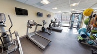 This fitness center is a comprehensively outfitted space designed to support a full range of workout routines. The room features durable rubber flooring throughout, a drop-panel ceiling with recessed fluorescent lighting, and warm neutral-toned walls. Large floor-to-ceiling windows along one side of the room flood the space with natural light while offering views of the surrounding neighborhood, creating an open and energizing atmosphere.

The cardio section is well-stocked with multiple treadmills, elliptical trainers, and a recumbent stationary bike, with wall-mounted flat-screen televisions positioned for easy viewing during workouts. A multi-function cable machine provides a versatile option for strength and resistance training. The free weights area includes a rack of kettlebells in varying sizes, medicine balls, stability balls, resistance bands, and foam rollers, all organized on dedicated storage racks.

A large wall-mounted mirror expands the sense of space and allows users to monitor their form. Posted fitness center rules near the entry door indicate a managed, community-oriented facility. Overall, the space is practical and efficiently organized, offering a balanced mix of cardio, strength, and functional training equipment in a clean and well-maintained environment.