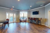 This single-floor multipurpose room is thoughtfully designed to serve as a communal gathering and event space. The interior is defined by rich wood-look tile flooring laid in a herringbone pattern throughout, complemented by soft gray walls and a distinctive teal-painted tray ceiling accented with white crown molding and recessed lighting that creates a polished, modern atmosphere.

The main area offers generous open floor space configured with multiple seating arrangements, including a standard dining table with upholstered leather chairs and a bar-height table surrounded by leather-and-metal barstools. A wall-mounted flat-screen television is positioned above the bar-height seating area, making the space well-suited for gatherings, screenings, or casual meetings.

Large windows and multiple sets of French doors allow abundant natural light to fill the room while providing direct access to an outdoor area, which appears to feature a pool and terrace. A full-wall mirror in one section of the room amplifies the sense of openness and reflects the natural light from the windows.

A built-in kitchenette along one wall features navy blue cabinetry with a light countertop, an integrated sink, and a compact refrigerator — providing convenient amenities for events and social functions. The overall design blends functional versatility with a warm, inviting aesthetic suited to a variety of uses.