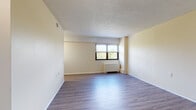 This single-floor unit offers a straightforward and functional layout designed to maximize usable living space. The main living area is open and generously proportioned, finished with smooth, light-toned hardwood-style flooring that runs throughout. Warm cream-colored walls reflect natural light, contributing to a bright and airy atmosphere. A large window fitted with horizontal blinds overlooks a verdant outdoor setting, bringing greenery and daylight into the space. Beneath the window sits a white radiator heating unit, providing a classic and efficient heating solution. The ceiling features a textured popcorn finish, adding subtle depth overhead. A dedicated entryway provides a clear transition into the main living space, complete with a sturdy door and a functional corridor. The bathroom is tiled from floor to surround, housing a full-sized soaking tub with chrome fixtures and grab bar support. The overall floorplan, as seen in the dollhouse view, is compact yet well-organized, with the bathroom and living area clearly delineated. Clean white trim and baseboards frame each room, lending a tidy, well-maintained appearance throughout the unit.