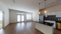 This single-floor unit is designed with an open-concept layout that seamlessly connects the living area and kitchen. The living space features warm hardwood-style flooring and receives generous natural light through large windows and a set of French doors, which open to enhance the airy, expansive feel of the area. A dedicated dining or entry zone transitions naturally into the kitchen, where tall ceilings draw the eye upward. The kitchen is well-appointed with white cabinetry, dark granite countertops, a tiled backsplash in a cool blue-grey tone, and stainless steel appliances including a refrigerator with ice maker, microwave, dishwasher, and a gas range. A central island with matching dark countertops provides additional workspace and storage, while pendant lighting and a track light fixture add focused illumination. The bedrooms are carpeted and feature ceiling fans, natural light from double or triple windows with blinds, and neutral wall tones. Both bathrooms include tile flooring, a tub/shower combination with white tile surround, a vanity with under-sink storage, and clean, functional fixtures throughout.