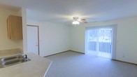 This single-floor space offers a straightforward and functional layout centered around a carpeted living area. The room features light grey carpeting that extends throughout, providing a neutral and comfortable foundation. A ceiling fan with an integrated light fixture is mounted at the center of the ceiling, offering both air circulation and ambient lighting. Along one wall, vertical blinds cover a large window or sliding door, allowing natural light to filter into the space while maintaining privacy. The white walls and light-colored ceiling contribute to a bright, open atmosphere. A standard interior door with wood trim is visible in the corner of the room, leading to an adjacent area. Baseboards frame the perimeter of the room cleanly. A partial countertop or surface is visible in the foreground, suggesting proximity to a kitchen or utility area. Overall, the space presents a clean, move-in ready environment with modest but practical finishes suited for everyday living.