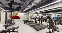 This fitness facility is designed to accommodate a wide range of workout disciplines within a single, thoughtfully organized floor. The main exercise area features an extensive selection of cardio equipment, including treadmills, stair climbers, and stationary bikes, each outfitted with individual digital screens for on-demand programming. A dedicated free weights section lines one wall, stocked with a full rack of dumbbells alongside adjustable benches and a cable machine. Large floor-to-ceiling mirrors are strategically positioned throughout to visually expand the space and support proper form during training.

The facility also includes a separate studio room, distinguished by a dark painted ceiling, built-in ballet barre along one wall, a gold-framed mirror, and a warm timber slatted accent panel. This versatile room is equipped with yoga mats and small weights, making it suitable for barre, yoga, stretching, or guided classes.

Throughout the main space, warm timber millwork cabinetry provides storage for rolled towels and houses a water station, offering a refined, hospitality-inspired touch. The overall aesthetic balances industrial elements—exposed black ductwork, metal beams, and linear LED strip lighting—with clean white floors and walls, creating a professional yet inviting atmosphere conducive to focused training.