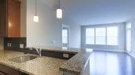 This single-floor residence showcases a thoughtfully designed open-concept layout that seamlessly connects the kitchen and living areas. The kitchen is a standout feature, boasting expansive granite countertops with rich, varied patterning that serve as both a functional workspace and a visual centerpiece. A deep undermount sink with a modern faucet adds to the kitchen's practicality, while stylish cylindrical pendant lights hang overhead, providing warm, focused illumination. The kitchen island extends generously into the space, offering additional seating and prep area, and is accented by a dark tile backsplash that contrasts elegantly with the light granite surfaces. The open living area beyond is bathed in natural light, courtesy of large windows fitted with blinds that allow for adjustable brightness throughout the day. The neutral, soft-toned walls and light flooring contribute to a clean, airy ambiance throughout. A doorway visible from the main living space leads to additional areas of the residence, reinforcing the home's flowing, connected layout. The overall design reflects a contemporary aesthetic, combining functional features with modern finishes to create a bright and inviting living environment.