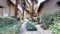 This single-floor residence is set within a thoughtfully landscaped community that immediately impresses with its natural surroundings. Tall, mature trees tower above the property, providing generous shade and a sense of established character throughout the grounds. Winding concrete pathways meander through well-tended garden beds filled with a diverse mix of shrubs, ground cover, and flowering plants, creating a serene, park-like atmosphere from the moment of arrival.

The exterior of the building features a neutral stucco finish complemented by wood-accented balconies and trim, reflecting a classic mid-century influenced architectural style. The unit's entry is particularly inviting, with a distinctive teal front door adorned with a decorative wreath, framed by lush, overhanging foliage that adds privacy and charm. The interplay of the natural landscaping with the built environment gives the property a secluded, tranquil feel despite being part of a larger complex.

The surrounding communal outdoor spaces are well-maintained, with layered plantings that provide year-round greenery and visual interest. The mature tree canopy filtering natural light across the pathways and garden areas enhances the overall ambiance, making the approach to the home a pleasant and scenic experience. This property offers a harmonious blend of comfortable residential living and lush natural beauty.
