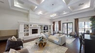 This single-story clubroom in Houston, TX is a refined communal space designed to impress. The main lounge area is defined by dramatic coffered ceilings that soar overhead, lending an airy, grand quality to the room. A floor-to-ceiling white built-in wall unit serves as the focal point, flanked by a classic fireplace with decorative tile surround and flanking open shelves displaying curated décor. Rich dark hardwood flooring grounds the space, contrasting elegantly with the bright white architectural millwork and paneled walls. Comfortable neutral-toned seating is arranged around a distinctive organic glass-and-wood coffee table, creating an inviting social atmosphere. Natural light filters in through large windows dressed in warm brown drapes, with French doors providing views of an outdoor area. A graceful arched doorway connects the lounge to additional areas of the clubroom. The kitchen area offers a striking contrast with dark charcoal walls, large-format white tile flooring, gray cabinetry, and sweeping granite countertops with a bold veining pattern. Sleek teardrop pendant lights illuminate the space, which is equipped with full-size appliances including a black refrigerator. Decorative round mirrors add a contemporary touch to this highly functional amenity space.