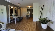 This single-floor unit presents an open-concept layout that seamlessly connects the living, dining, and kitchen areas into one cohesive space. Warm wood-look plank flooring extends throughout, providing a consistent and inviting foundation underfoot.

The living area accommodates a compact seating arrangement, while the adjacent dining space features a rectangular table with seating for several guests. A large mirror on the wall visually expands the area, enhancing the sense of openness. Neutral-toned walls, including a contrasting dark accent wall, add depth and character to the space.

The kitchen is defined by dark espresso cabinetry that reaches toward the ceiling, stainless steel appliances including a refrigerator, and a central island with bar-height seating — ideal for casual dining or food preparation. Recessed and flush-mount ceiling lighting provides consistent illumination throughout the space, complemented by a track light fixture in the living area. A doorway with a louvered door is visible near the entrance, suggesting additional storage or utility access. Overall, the space conveys a modern, functional, and well-appointed design aesthetic.