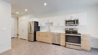 This single-floor unit showcases a thoughtfully designed, modern interior with a clean, neutral palette throughout. The kitchen is a standout feature, outfitted with two-tone cabinetry — warm natural wood lower cabinets paired with crisp white upper cabinets — complemented by a white hexagonal tile backsplash and light quartz countertops. A full suite of stainless steel appliances includes a French door refrigerator, electric range, over-the-range microwave, and dishwasher. A built-in countertop extension provides additional workspace or casual dining space.

The open living area is bright and versatile, featuring recessed lighting, neutral luxury vinyl flooring, and white walls that create a clean, airy feel. A glass-paneled exterior door and adjacent window allow natural light to filter into the space, enhancing its open character.

The bathroom is finished in a minimalist style, with a glass-enclosed shower/tub combination, brushed nickel grab bars, and matching flooring that carries the cohesive aesthetic throughout. Closet storage is accessible from the main living area. The overall design prioritizes functionality and modern simplicity, offering a well-appointed, move-in-ready space.