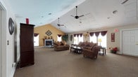 Located at 8799 Old Colony Way in Alexandria, Virginia, this single-story community clubhouse offers a welcoming array of amenities designed for both relaxation and recreation. The interior centers around a generously proportioned main lounge featuring soaring vaulted ceilings accented by ceiling fans and decorative panel detailing. Warm golden walls, plush upholstered seating, rich dark wood furnishings, and a traditional fireplace create an inviting and comfortable gathering atmosphere. Multiple windows dressed with deep burgundy drapes allow natural light to fill the space. An adjacent foyer area with vaulted ceilings, a large decorative mirror, and elegant console table provides a gracious entry point connecting the interior rooms through classic double doors. The clubhouse also includes a fully functional kitchen equipped with dark wood cabinetry, granite-style countertops, and white appliances, making it well-suited for events and gatherings. Outdoors, the facility boasts a large swimming pool surrounded by a spacious deck with lounge seating, complemented by a dedicated pool house with changing facilities. A set of well-maintained tennis courts rounds out the outdoor amenities, set against a backdrop of mature trees and residential buildings.