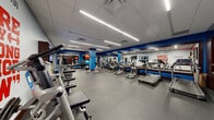 This fitness center is a comprehensively outfitted workout facility designed to accommodate a wide variety of exercise needs. The space is defined by a bold blue and white color scheme, with vibrant accent walls and motivational phrases such as "No Excuses," "Be Active, Be Healthy, Be Happy," and "Sore Today, Strong Tomorrow" displayed prominently throughout, creating an energizing atmosphere.

The facility features a dedicated cardio zone lined with treadmills, elliptical trainers, and stationary bikes, all positioned in front of large wall-to-wall mirrors that visually expand the space and allow users to monitor their form. Flat-screen televisions are mounted at multiple points throughout the room, ensuring entertainment is accessible from virtually any workout station.

The strength training area includes a well-stocked dumbbell rack with a full range of free weights, adjustable benches, stability balls, and a foam roller for stretching and recovery. Cable machines and multi-functional strength units are also present, offering options for both isolation and compound movements.

The flooring throughout consists of durable rubber gym matting, providing safety and comfort underfoot. The drop ceiling features recessed linear LED lighting that ensures bright, even illumination across the entire floor. Locker storage and additional amenities are accessible from an area near the rear of the facility, rounding out this practical and well-designed fitness space.