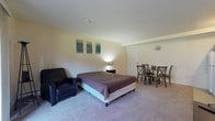 This single-floor studio presents an efficiently organized open-concept layout, seamlessly integrating the living, sleeping, and dining functions within one generously proportioned room. The main living area is finished with wall-to-wall carpeting in a neutral tone, contributing to a warm and cohesive atmosphere throughout. A full-sized bed serves as the central anchor of the space, complemented by a black leather armchair and a compact dining set with cross-back chairs — all arranged to maximize usability within the open floor plan. A wall-mounted flat-screen television and a small media console add a functional entertainment element to the room. A sliding glass door floods the space with natural light and provides direct access to an outdoor balcony, enhancing the sense of openness.

The kitchenette, tucked into one corner of the unit, features warm wood cabinetry, a gas range with overhead ventilation, a refrigerator, a sink with dark countertops, and tile flooring — providing all essential cooking amenities in a compact footprint. The bathroom is finished with classic white square-tile walls, and includes a tub-shower combination, toilet, and a single vanity sink topped with a mirrored medicine cabinet, offering clean and functional design.
