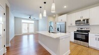 This well-designed 2-bedroom, 2-bathroom single-floor residence offers a thoughtful layout with modern finishes throughout. The open-concept kitchen and living area serves as the heart of the home, anchored by a large center island with a striking marble-style countertop, matte black faucet, and an undermount sink. White shaker-style cabinetry lines the kitchen walls, complemented by stainless steel appliances including a French door refrigerator, range, and over-the-range microwave. A trio of pendant lights illuminates the island, adding a contemporary touch. Warm hardwood-style flooring flows throughout the main living areas, while the living room features a ceiling fan and a set of French doors with transom window above, inviting natural light and outdoor access. The bedrooms are carpeted for comfort, each equipped with a ceiling fan and windows fitted with blinds. The primary bedroom includes dedicated closet space. Both bathrooms are finished with white cabinetry, marble-style countertops, matte black hardware, and hardwood-style flooring, with the primary bath offering a vanity with a large mirror and direct access to a walk-in closet. The overall aesthetic is clean, bright, and contemporary.