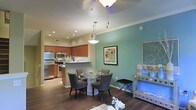 This two-floor residence showcases a thoughtfully designed open-concept living space that seamlessly connects the kitchen and dining areas. The kitchen is outfitted with warm wood cabinetry, stainless steel appliances including a refrigerator, microwave, and range, and is anchored by a peninsula that provides additional prep space and a natural transition into the dining area. Recessed lighting throughout the kitchen ensures ample illumination, while a pendant light adds a stylish focal point above the peninsula.

The dining area is distinguished by a teal accent wall that adds a bold splash of color against the neutral tones of the surrounding space. A round glass-top dining table surrounded by upholstered chairs in charcoal grey creates an inviting setting. A modern pendant light hangs overhead, complementing the ceiling fan nearby. Rich, dark hardwood flooring runs throughout, lending warmth and continuity to the space. Crown molding along the ceiling adds a refined architectural detail. A decorative console table along the wall provides additional display and storage space, completing the polished aesthetic of this well-appointed home.