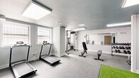 Located at 1228 Spruce Street in Philadelphia's vibrant Washington Square West neighborhood, this well-appointed fitness center offers a thoughtfully organized exercise space across a single floor. The room is finished with durable speckled gray flooring throughout, complemented by crisp white walls adorned with subtle sage green geometric accents that lend a modern, energizing aesthetic. A dark teal ceiling adds a bold design contrast, while large flush LED panel lights ensure bright, even illumination across the entire space. Natural light filters in through windows along one wall, enhancing the open feel of the room. The cardio zone features multiple treadmills, while the strength area is equipped with a multi-function cable machine, an adjustable weight bench, and a rack of dumbbells in varied sizes. A dedicated turf section provides a versatile surface for functional training, stretching, or bodyweight exercises. A wall-mounted yoga mat and a water dispenser add practical convenience. A full-wall mirror visually expands the space and supports form-focused training. An adjoining doorway leads to an adjacent utility or changing area, rounding out the functionality of this compact but comprehensive fitness facility.