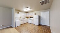 This 1-bedroom, 1-bathroom single-floor unit offers a practical and comfortable layout designed for efficient living. The kitchen is a functional highlight of the space, featuring white cabinetry, a full suite of white appliances including a refrigerator and electric range, a double-basin sink, and wood-look vinyl flooring. A stacked washer and dryer unit is conveniently integrated directly into the kitchen area, adding exceptional in-unit utility. The kitchen flows naturally into the entry and dining areas, which share the same durable wood-look flooring and are bathed in neutral, warm-toned walls throughout.

The living room is generously sized with soft carpeting underfoot and benefits from natural light through multiple windows fitted with blinds, as well as a glass-paneled exterior door that further brightens the space. The bedroom offers a comfortable footprint with plush carpeting, a window with blinds providing natural light, and access to a dedicated closet equipped with wire shelving for organized storage.

The bathroom features wood-look vinyl flooring consistent with the rest of the unit, a vanity with countertop sink, a toilet, and grab bars, suggesting accessibility-conscious design. Clean white trim and beige-toned walls provide a cohesive, neutral aesthetic throughout the entire space.