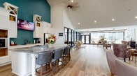 This single-story community clubhouse is thoughtfully designed to offer a variety of functional and inviting spaces under one roof. The interior is unified throughout by wide-plank light hardwood flooring and crisp white walls, creating a bright, cohesive aesthetic.

The kitchen area is a standout feature, showcasing soaring vaulted ceilings, bold teal accent walls, and white shaker-style cabinetry extending to the ceiling. A large marble-topped island with bar seating serves as a central gathering point, complemented by stainless steel appliances, a wall-mounted television, and abundant counter space. An adjacent workstation area provides a dedicated business center.

The office area features dramatic vaulted ceilings with an arched transom window above French doors, flooding the space with natural light. A chandelier adds an elegant focal point, while a formal desk arrangement supports professional meetings or leasing consultations.

A generous lounge area offers plush seating around a central coffee table, with large mirrored doors leading directly into a well-equipped fitness center. Throughout, dark-framed architectural details contrast cleanly against light walls.

A glass-walled sunroom at the rear of the space is bathed in natural light from expansive windows and French doors that open to an outdoor area, providing a seamless indoor-outdoor connection ideal for relaxing or socializing.