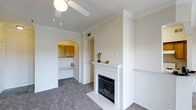 This single-floor residence is thoughtfully arranged to maximize both functionality and comfort. The living area serves as the centerpiece of the space, featuring a classic white-mantled fireplace with a tile surround that acts as a natural focal point. Crown molding lines the ceiling, adding a refined architectural detail, while a ceiling fan provides practical comfort. Soft carpet flooring extends throughout the main living and bedroom areas, contributing to a warm, cohesive aesthetic.

A distinctive arched entryway connects the living area to the kitchen, providing an elegant transition between spaces. The kitchen features wooden cabinetry and a lighter countertop surface, offering ample storage and workspace. A built-in counter or desk area is visible near the kitchen, adding versatile utility to the layout.

The floor plan, as seen from above, reveals a well-organized arrangement of rooms branching from a central corridor, including what appear to be multiple bedrooms and a bathroom with a checkered tile detail. Each room is finished with consistent neutral tones throughout, creating a clean, bright, and inviting atmosphere across the entire space.