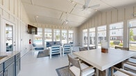 This single-story amenity clubhouse is designed as a light-filled gathering space with a coastal-inspired aesthetic. The vaulted, paneled ceiling rises prominently overhead, enhancing the sense of openness throughout the room, while dual ceiling fans provide consistent airflow across the entire space.

Floor-to-ceiling windows line multiple walls, flooding the interior with abundant natural light and offering unobstructed views of the surrounding outdoor pool area and landscaping. The white board-and-batten wall paneling reinforces a clean, coastal design language consistent throughout.

The space is functionally divided into distinct zones. A lounge area features white-framed furniture with blue upholstered cushions arranged around a low coffee table atop a patterned area rug. Adjacent to this is a dining zone with a neutral-toned rectangular table and woven accent chairs, suitable for group gatherings. A wall-mounted television is positioned near the lounge seating.

A kitchenette along one wall includes gray cabinetry with granite countertops, a stainless steel refrigerator, microwave, and sink, providing practical refreshment capabilities. A separate exterior door offers direct outdoor access. The polished concrete flooring ties all areas together with a durable, cohesive finish.
