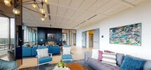 Located at 90 Neilson St in New Brunswick, NJ, this single-floor communal space offers a refined and contemporary atmosphere designed for comfort and social interaction. The lounge is anchored by expansive floor-to-ceiling glass walls that flood the interior with natural light and frame sweeping views of the surrounding New Brunswick skyline and cityscape.

The seating areas are thoughtfully arranged, featuring a mix of plush sofas, accent chairs in bold blue tones, and patterned area rugs that add texture and warmth to the polished tile flooring. A sleek, dark-paneled feature wall houses a large flat-screen display and a modern linear fireplace, serving as a striking focal point within the space.

Stylish pendant lighting with an artistic, multi-globe design complements the recessed ceiling lights, contributing to a layered and inviting ambiance. Curated original artwork is displayed prominently along the crisp white corridor walls, giving the space a gallery-like quality as visitors move through it.

Access to an outdoor terrace is provided through full-height glass doors, further connecting the interior to the outdoors. The corridor features institutional-grade finishes with clean lines, automated doors, and efficient layout, reflecting a well-designed professional or residential amenity space built for everyday use.