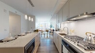 This thoughtfully designed single-floor residence combines modern aesthetics with functional living. The open-concept kitchen serves as the heart of the home, featuring soft gray shaker-style cabinetry, quartz countertops with a chevron-patterned tile backsplash, stainless steel appliances including a gas range and built-in microwave, and a generous island with pendant lighting overhead. Warm wood-tone flooring runs throughout, lending continuity and a welcoming feel across all areas. The kitchen flows seamlessly into a dining area and an inviting living room, both bathed in abundant natural light from expansive floor-to-ceiling windows that frame views of the surrounding urban skyline. The living area offers a relaxed yet stylish atmosphere, with ample room for comfortable seating arrangements. The bedroom continues the bright, airy theme with its own large corner window showcasing open city views, creating a serene retreat. The bathroom is clean and contemporary, finished in crisp white with light wood-look tile flooring, a full soaking tub with shower, and a white vanity with under-cabinet storage. Throughout the space, a consistent palette of whites, grays, and warm natural tones creates a cohesive, modern ambiance that feels both refined and livable.