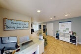 Located at 313 Lincoln Avenue in College Station, Texas, this single-floor leasing office for University Square Apartments offers a compact yet efficiently organized environment designed to welcome prospective and current residents. The main reception area greets visitors with a sleek white front desk, recessed lighting, and warm tile flooring throughout. A cheerful "Welcome to your new home" sign sets a friendly tone, while apartment floor plan displays and informational materials line the walls, providing prospective residents with a clear overview of available unit styles. Natural light filters in through a sliding glass door dressed with patterned curtains, brightening the space. The office area features a dedicated workstation with a desktop computer, whiteboard, and printer, supporting day-to-day leasing operations. Decorative touches such as potted plants, ornamental grasses, and a bold "HOWDY" display add personality and reflect the Aggie spirit of the College Station community. The kitchen area is outfitted with dark wood-grain cabinetry, granite-style countertops, a microwave, mini-refrigerator, and a coffee station, offering staff and visitors a comfortable amenity. The overall ambiance is clean, organized, and inviting — a practical and pleasant first point of contact for the community.