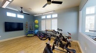 This fitness room offers a thoughtfully designed workout space finished with light hardwood flooring and soft blue walls that create a calm, energizing atmosphere. The room benefits from generous natural light streaming through a row of large windows fitted with white horizontal blinds, complemented by clerestory windows above that further brighten the space. Ceiling fans ensure comfortable airflow during workouts.

The room is outfitted with multiple spin bikes positioned toward the center, providing ample space for cycling sessions. A tiered kettlebell rack holds a range of weights for strength training, while a dedicated storage rack organizes resistance bands, medicine balls in various sizes, and exercise mats, keeping the space tidy and functional. A wall-mounted television adds an entertainment or instructional element to workouts.

The open layout allows for versatile use, accommodating both cardio and functional training activities. The combination of durable flooring, strategic equipment placement, and abundant natural light makes this fitness space both practical and inviting for a variety of exercise routines.