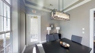 This single-floor space showcases a refined and contemporary interior design with carefully considered details throughout. The standout dining area draws immediate attention with its striking rectangular pendant light fixture, featuring a dark metal frame and warm ambient panels that cast a welcoming glow over the dark-toned dining table below. The tray ceiling adds architectural depth and dimension to the room, while crown molding provides a polished, finished look along the upper walls. Soft gray walls create a neutral, sophisticated backdrop that complements the dark furnishings, including a sleek sideboard adorned with matching table lamps and a decorative sunburst mirror that adds an artistic focal point. A classic French door with multi-pane glass panels invites abundant natural light into the space, offering a view to the exterior and enhancing the overall sense of openness. Gray upholstered dining chairs contribute to the cohesive, modern aesthetic. The combination of layered lighting, architectural ceiling details, and thoughtfully curated décor creates an ambiance that is both elegant and inviting, making this space well-suited for gatherings and daily use alike.