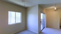 This single-floor residence offers a clean, well-maintained living environment with a straightforward and functional layout. The interior is finished with neutral-toned walls and wall-to-wall carpeting throughout, creating a warm and cohesive atmosphere across the space.

The main living area is defined by its notably tall, vaulted ceilings, which lend an open and airy quality to the room. A decorative pendant light fixture hangs from the ceiling, adding a subtle design element to the otherwise understated space. Natural light filters in generously through a large window on the side wall, offering views of surrounding greenery that contribute to a calm and pleasant ambiance.

A sliding glass door provides access to an adjacent outdoor area — likely a patio or balcony — allowing for seamless indoor-outdoor connectivity and additional natural light. The partially visible exterior through the glass door suggests a bright, open outdoor space.

The interior walls are smooth and painted in a soft, neutral tone, complementing the light-colored carpeting and contributing to the overall sense of cleanliness and simplicity. The space presents a move-in-ready condition with a modest yet comfortable aesthetic, well-suited for those seeking a bright, quiet, and functional single-level living environment.
