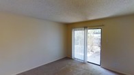 This single-story space features a modest, functional layout with a clean and straightforward design. The main living area is finished with wall-to-wall carpeting in a neutral tone, providing a comfortable underfoot feel throughout the room. The walls are painted in a soft, warm beige, contributing to an understated and practical ambiance.

A textured popcorn ceiling adds a classic touch to the interior, a common architectural detail that also aids in sound absorption. The room is illuminated by natural light streaming in through a full-height sliding glass door, which serves as the focal point of the space. Fitted with vertical blinds, the slider allows for adjustable light control and privacy.

The sliding glass door opens directly to an outdoor patio area, visible through the glass and framed by a wooden fence and surrounding greenery. This connection to the outdoors enhances the sense of openness within the room, making the space feel more expansive than its footprint suggests. Overall, the space offers a practical and comfortable environment suited to everyday living.