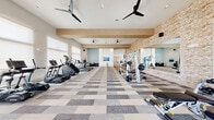 Located at 150 Inspiration Dr in Hudson Oaks, TX, this fitness center offers a generously sized, single-floor workout facility designed with both function and aesthetics in mind. The space features an open layout with high ceilings accented by warm wood beams and prominent stacked stone accent walls that add a refined, contemporary character to the interior.

The flooring throughout is a patterned modular carpet tile in neutral tones of grey, beige, and cream, providing both durability and visual appeal. Ceiling fans are strategically placed to promote airflow across the workout floor. Large windows along multiple walls flood the space with natural light, while also offering views to the exterior.

The cardio zone is well-appointed with a row of treadmills, ellipticals, and stair climbers from the TRUE Fitness brand, each equipped with individual display screens. A wall-mounted flat-screen TV is positioned to keep members entertained during their sessions. A dedicated cycling area features stationary and spin bikes positioned near bright, oversized windows.

The strength training area is equally impressive, with a full complement of cable machines, weight benches, and a neatly organized dumbbell rack. Floor-to-ceiling mirrors line the stone accent wall, visually expanding the space and supporting proper form during exercises. The overall ambiance is clean, modern, and inviting—ideal for a focused and comfortable workout experience.