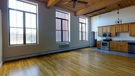 This single-floor loft-style space showcases a striking blend of industrial and warm design elements that come together to create a character-rich interior. The standout feature is the exposed wooden beam ceiling, which adds a rustic, authentic charm and a sense of architectural history to the space. A ceiling fan is centrally mounted, enhancing both functionality and visual interest overhead.

The open-concept layout allows the main living area to flow seamlessly into the kitchen, maximizing the sense of spaciousness. Large, multi-pane industrial-style windows line the walls, framing views of the surrounding urban environment while flooding the interior with generous amounts of natural light. The windows' bold black frames add a contemporary contrast to the warm wood tones throughout.

Underfoot, light-toned hardwood flooring runs throughout the space, contributing to its airy and inviting ambiance. The kitchen area features warm wood cabinetry and is equipped with stainless steel appliances, including a range and microwave, offering a practical and stylish cooking space. A wall-mounted PTAC unit provides climate control along the main living wall.

Overall, the space exudes an open, loft-inspired atmosphere with a thoughtful combination of raw industrial elements and comfortable, modern finishes.