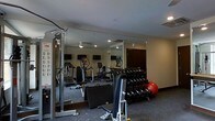 This fitness center is a fully outfitted exercise facility designed to accommodate a wide range of workout routines. The space features a rubberized flooring surface that provides both durability and safety during training sessions. A large floor-to-ceiling mirror spans one wall, visually expanding the room while allowing users to monitor their form during exercises.

The cardio section is equipped with a selection of machines including treadmills, elliptical trainers, and stationary bikes, each fitted with individual display monitors for entertainment or performance tracking. A dedicated free weights area includes a multi-tier dumbbell rack stocked with a full range of weights, alongside a stability ball for functional training.

Strength training is further supported by a multi-station cable and pulley machine, as well as an adjustable weight bench, offering versatility for users at varying fitness levels. Recessed lighting throughout the ceiling provides consistent, even illumination across the entire workout floor. A dark wood door provides access to and from the facility. The overall ambiance is clean, functional, and purposefully designed to create a focused and motivating environment for fitness enthusiasts.