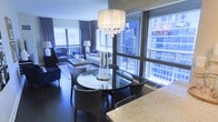 This stylish single-floor residence offers an open-concept living and dining area that seamlessly blends elegance with urban sophistication. Floor-to-ceiling windows wrap the space, flooding the interior with natural light while framing dramatic city skyline views that serve as a striking backdrop. The dining area is anchored by a round glass-top table surrounded by sleek dark chairs, illuminated overhead by a stunning crystal-accented drum chandelier that adds a glamorous focal point to the space. Adjacent to the dining area, a granite-topped kitchen counter transitions smoothly into the living space, hinting at a well-appointed culinary area. The living room features a carefully curated arrangement of neutral-toned sofas and deep navy accent chairs, layered over warm hardwood flooring that runs throughout. Soft ambient lighting complements the cool blue tones cast through the expansive windows. Decorative candle holders and refined accessories add texture and warmth to the overall aesthetic. The cohesive color palette of navy, grey, and warm neutrals throughout creates a polished, contemporary ambiance befitting its prominent urban setting.