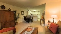 This single-floor residence showcases an open-concept design that seamlessly connects the living, dining, and kitchen areas into one cohesive, flowing space. The living area, positioned in the foreground, features plush carpeted flooring in a warm neutral tone and is furnished with a wooden coffee table, offering a relaxed and welcoming atmosphere. A tall decorative plant adds a natural touch alongside a classic wooden armoire.

The dining area sits centrally within the space, anchored by a dark wood dining table and matching chairs, creating a defined zone within the open layout. Decorative accents on the table contribute to an inviting, lived-in feel.

The kitchen, visible in the background, features white cabinetry on both upper and lower levels, providing ample storage. A peninsula counter separates the kitchen from the dining area, and standard appliances including a range and oven are integrated into the layout. Recessed and overhead lighting illuminate the kitchen area, while the overall space benefits from warm ambient lighting throughout. The textured ceiling and neutral wall tones tie the entire open-plan area together cohesively.
