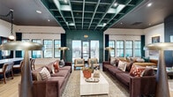Located at 214 Kingsmeadow Cir in Belville, NC, this thoughtfully designed single-floor community space offers a generous footprint across a seamlessly connected layout. The centerpiece is a dramatic main lounge anchored by a striking teal coffered ceiling, light hardwood flooring, and an array of curated seating arrangements including plush sofas, accent chairs, and statement floor lamps. Large black-framed windows flood the space with natural light and provide direct views of the outdoor pool area.

A secondary lounge area features a tiled fireplace wall with a mounted TV, cozy caramel-toned seating, and intimate scale—ideal for relaxed gatherings. A third sitting room showcases an eclectic gallery wall, textured wallcoverings, and a bold arched mirror that adds artistic flair.

The full kitchen is outfitted with sleek dark cabinetry, warm terracotta tile accents, chain-hung pendant lighting, and an elongated island with bar seating—well-suited for entertaining. A dedicated business center provides a granite-topped workstation with desktop computers, patterned wall art, and a built-in banquette beneath expansive windows. Throughout, the space maintains a cohesive, design-forward aesthetic with rich color palettes, curated artwork, and abundant natural light.