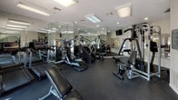 This fitness center is a comprehensively outfitted, single-floor facility designed to support a full range of workout routines. The space features durable dark rubber flooring throughout, providing both safety and functionality for a variety of exercises. Bright flush-mounted ceiling panels and recessed lighting ensure the room is well-lit at all times, complemented by natural light streaming in through windows fitted with horizontal blinds and a set of glass double doors that open to the exterior.

Floor-to-ceiling mirrors line the walls, visually expanding the space and allowing users to monitor their form during workouts. The cardio zone is well-represented with multiple treadmills, elliptical trainers, and stair-climber machines. A multi-station cable machine provides versatile options for strength training, while a dedicated free weights area features a full dumbbell rack alongside an adjustable workout bench. A mounted flat-screen television is positioned for easy viewing during exercise sessions.

The neutral gray walls and clean, uncluttered layout contribute to an organized and motivating atmosphere. Informational postings on the walls offer guidance on facility rules and workout tips. The overall design is practical and efficient, making this fitness center a functional and accessible resource for all fitness levels.