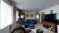 This multi-floor home spans three levels, offering a variety of living spaces designed for everyday comfort and functionality. The main living area is a carpeted room that provides a warm and inviting atmosphere, centered around a generously sized seating arrangement featuring a plush sectional sofa with accent pillows and a reclining chair. Natural light filters in through a window dressed with patterned curtains, brightening the space without overwhelming it. Wooden cabinetry and open bookshelves line the walls, offering both decorative display and practical storage options. Framed artwork adorns the neutral-toned walls, adding character to the room. A large flat-screen television is positioned on a wooden media console, making the space well-suited for relaxing and entertainment. The textured ceiling adds a classic touch to the overall aesthetic. A doorway visible from the living area hints at additional rooms beyond, suggesting a well-connected floor plan. With its understated, traditional design and thoughtful layout, this home provides a comfortable and livable environment spread across all three floors.