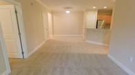 This single-floor unit features an open-concept design that seamlessly connects the main living area to the kitchen. The living space is finished with soft, plush wall-to-wall carpeting in a warm neutral tone, complemented by beige walls and crisp white baseboards that create a clean, cohesive aesthetic throughout. Recessed lighting is paired with a central ceiling-mounted light fixture, ensuring the space is well-lit. Crown molding adds a subtle architectural detail along the ceiling line, lending a polished finish to the interior.

The kitchen is positioned to the side of the main living area and is partially separated by a raised breakfast bar countertop with a dark granite surface, offering both a casual dining option and a natural visual divider. Warm-toned wood cabinetry lines the upper portion of the kitchen, while a stainless steel refrigerator anchors the space and adds a modern contrast to the warm finishes. Interior doors provide access to additional rooms, and the overall layout is practical and functional, making efficient use of the available space.