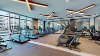 Located at 1 Midtown Bridge Approach in Hackensack, NJ, this contemporary fitness center is thoughtfully designed to accommodate a full range of workout needs across distinct functional zones. The space is characterized by a striking ceiling design featuring geometric, cross-pattern LED lighting fixtures with warm-toned accents set against a clean white backdrop, lending a sleek, upscale ambiance throughout.

The primary cardio area is expansive, lined with rows of treadmills, ellipticals, and rowing machines positioned along floor-to-ceiling windows that flood the space with natural light and offer views of the surrounding streetscape. Large mirrors along the walls visually extend the space and enhance the open feel. Durable, patterned carpet tile flooring grounds the cardio zones.

A dedicated strength training area features cable machines, weight racks, benches, and free weights, all set against mirrored walls and accented by a bold graphic mural in navy, white, and gold—adding a distinctive design element.

A separate studio room offers light hardwood flooring, full-length mirrors, a ballet barre, a large display screen, a punching bag, and spin bikes—suitable for group classes or individual training. The studio also benefits from natural light via street-facing windows, completing this well-appointed facility.