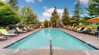 Located in Hillsboro, Oregon, this well-appointed outdoor amenity area centers around a generously sized swimming pool with crystal-clear blue water and a gradual entry staircase. The expansive pool deck is finished in a textured aggregate surface and is lined with multiple lounge chairs, providing ample space for sunbathing and relaxation. The pool is enclosed by a black metal fence, framed by mature evergreen and deciduous trees that lend a sense of privacy and natural beauty to the setting. At one end of the pool area, a dedicated outdoor living zone features a cozy fire pit with surrounding seating, perfect for cooler Pacific Northwest evenings. An outdoor kitchen and bar area, complete with a wood-fired pizza oven and covered patio seating with vibrant yellow umbrellas, offers an ideal setting for entertaining. The adjacent single-story clubhouse, with a pitched roof and large windows, opens directly onto the pool deck, providing seamless indoor-outdoor access. Solar panels are visible atop the clubhouse roof, reflecting an energy-conscious design. A decorative pergola structure anchors one corner of the grounds, adding architectural interest. The overall atmosphere is resort-like, combining functional amenities with lush, manicured landscaping throughout.