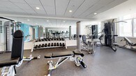 This fitness center offers a comprehensive workout environment designed to accommodate a wide range of exercise needs. The space is thoughtfully organized into distinct functional zones, all unified by durable speckled rubber flooring, a drop-panel ceiling with recessed lighting, and large mirrors that visually expand the room throughout.

The strength training area is equipped with a full dumbbell rack ranging across multiple weight increments, an adjustable weight bench, and a multi-station cable machine suitable for a variety of resistance exercises. Nearby, additional selectorized machines provide targeted muscle training options.

The cardio section features multiple treadmills, elliptical cross-trainers, and a recumbent-style cardio machine. Floor-to-ceiling windows line one wall, flooding this area with natural light and offering open outdoor views, creating an airy, motivating atmosphere during workouts.

A utility area includes dual water fountains mounted on a bold teal accent wall, along with restroom access and a clearly marked emergency exit. The neutral gray and white color palette, punctuated by the teal accent, gives the space a clean, modern aesthetic that is both functional and visually inviting.