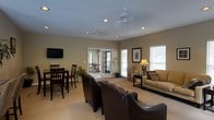 This single-floor space offers an open-concept layout that seamlessly integrates a living area and dining space. The living room is anchored by a seating arrangement featuring dark leather armchairs and a neutral-toned sofa, complemented by a wall-mounted flat-screen television. Multiple ceiling fans are distributed throughout, contributing to efficient air circulation across the expansive space.

Natural light flows generously through large windows fitted with horizontal blinds, as well as through a set of French doors that open to an outdoor patio area, providing views of the exterior. The warm beige carpeting underfoot lends a cohesive, comfortable feel to the interior.

The dining area is positioned adjacent to the living space and accommodates a dark wood table paired with matching bar-height chairs, making it suitable for casual meals. Decorative touches, including framed wall art, a table lamp, and potted plants near the French doors, add warmth and character to the overall ambiance. Recessed lighting in the ceiling provides additional illumination, enhancing the bright and welcoming atmosphere of the space.