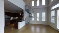 This two-story space makes an immediate impression with its soaring double-height ceilings and an eye-catching steel spiral staircase that serves as the central architectural focal point, gracefully connecting the two levels. The open-concept main floor seamlessly integrates the living area and kitchen, creating a fluid and functional layout. Large vertical windows along the upper portion of the walls flood the interior with natural light, while additional windows on the main level offer views of the surrounding area and further brighten the space.

The kitchen is outfitted with rich, dark wood cabinetry, stainless steel appliances including a built-in microwave and range, and a dark stone countertop that wraps around a peninsula with breakfast bar seating. Pendant lighting hangs above the kitchen area, complementing the track lighting overhead for layered illumination throughout.

The main living area features polished concrete flooring in a warm neutral tone, lending an industrial yet refined character to the space. Clean white walls throughout keep the interior feeling bright and open, while the overall design balances modern industrial elements with functional everyday living across both floors.