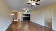 This single-story residence at Vineland Carriage Homes offers an open-concept layout that seamlessly connects the living area to the kitchen, creating a spacious and functional flow throughout the main living space. Rich hardwood-style flooring extends across the entire area, adding warmth and visual continuity to the interior.

The living area is generously sized, with ceiling fans providing both comfort and casual lighting. Neutral, warm-toned walls complement the flooring and contribute to a welcoming ambiance throughout the space.

The kitchen features dark cabinetry paired with black appliances, including a refrigerator and dishwasher, creating a sleek, cohesive aesthetic. A double-basin sink sits beneath the countertop, adding practicality to the well-appointed cooking space.

A hallway visible from the main living area leads further into the home, suggesting additional rooms beyond. Wall sconces along the corridor provide subtle accent lighting, enhancing the overall warmth of the interior. White baseboards throughout offer a clean contrast against the flooring and walls, giving the space a polished, finished appearance.
