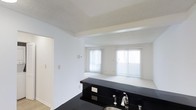 This single-floor space showcases a practical and visually striking open-plan design that connects the kitchen area directly to the living space. The kitchen is anchored by a bold black granite countertop with built-in electrical outlets, providing both aesthetic contrast and functional workspace. A stainless steel sink with a modern faucet adds a polished finish to the kitchen area.

A structural beam defines the transition between the kitchen and the adjoining living space, creating a subtle architectural division while maintaining an open, airy feel. The living area is finished with light-toned carpeting, contributing to a clean and neutral aesthetic throughout.

Two windows dressed with vertical blinds allow generous natural light to fill the living space, enhancing the bright, white-painted walls. The textured ceilings add a layer of visual depth to the otherwise minimalist interior palette.

A doorway off the kitchen provides access to an additional area of the unit, suggesting practical room separation within the single-floor layout. Overall, the space presents a straightforward, functional design with a notable contrast between the dark kitchen surfaces and the light-toned living area.