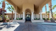 This single-story facility presents a striking Mediterranean-inspired exterior, defined by a grand arched entryway flanked by classical white columns set against warm stucco walls. Decorative stamped concrete pathways lead to double glass doors framed by lush landscaping, creating a welcoming and polished first impression.

Inside, the main lobby is designed with sophistication and comfort in mind. Soaring vaulted ceilings with crown molding establish a sense of grandeur, while a dramatic pendant chandelier — featuring cascading glass or crystal elements — serves as a stunning focal point overhead. The seating area is elegantly arranged with high-back upholstered wing chairs in muted blue-gray tones surrounding a round marble-top accent table, offering an inviting space for conversation and relaxation.

A sleek, floor-to-ceiling fireplace surround anchors one wall, complemented by a mounted flat-screen display above. The warm wood-toned flooring adds texture and warmth throughout. Beyond the lobby, additional gathering and activity rooms are visible, featuring teal accent walls and ample natural light, suggesting a variety of social and recreational uses. The overall ambiance is refined yet approachable, blending upscale design with a community-oriented atmosphere.