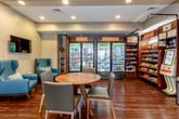 This single-floor amenity space is thoughtfully designed to serve multiple functions within a cohesive, well-appointed layout. Rich hardwood flooring runs throughout, anchoring the neutral gray walls and dark wood trim that define the overall aesthetic.

A fitness center is visible through a series of large, dark-framed glass windows and doors along one wall, offering a transparent view into a well-equipped gym with cardio machines, strength equipment, and ceiling fans. The open sightlines create a sense of connectivity between the workout area and the adjoining lounge space.

The lounge area features a round wooden dining table surrounded by gray upholstered chairs, alongside a teal wingback armchair paired with a gold accent table. A wall-mounted flat-screen television adds a functional entertainment element to this relaxed seating zone.

A self-service kiosk station is positioned near the entrance, providing convenient access to additional services. Adjacent to the lounge, a well-stocked convenience market area includes multiple glass-door refrigerated units filled with beverages, alongside shelving units displaying a variety of packaged snacks and grab-and-go items.

Recessed lighting throughout ensures consistent, even illumination across all areas of the space.